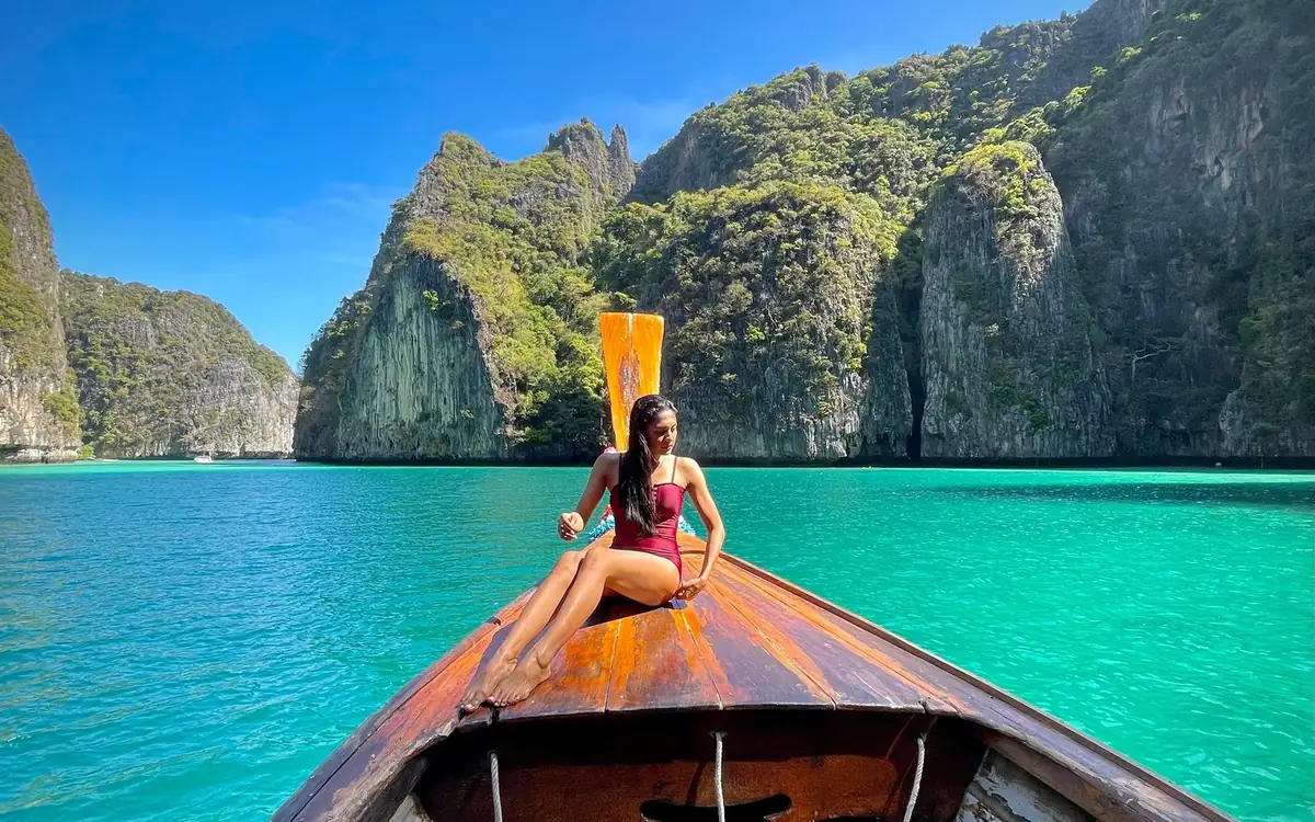 The wooden bow of a longtail boat with a woman sitting on it, in a turquoise lagoon below green limestone cliffs