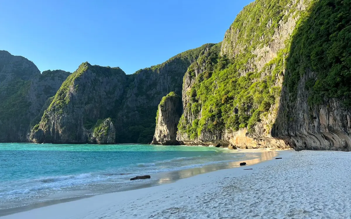 An empty white sand beach below steep green limestone cliffs, with nobody on it