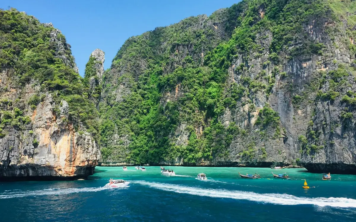 A speedboat under way in a lagoon between green limestone cliffs, with more boats moored along the far shore