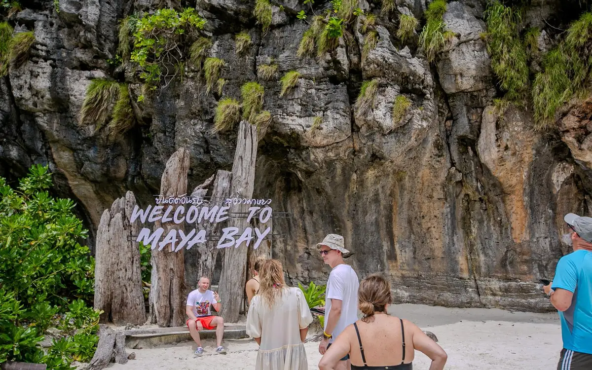 Visitors standing on the sand in front of a Welcome to Maya Bay sign below a cliff